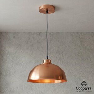 May include: A copper-colored pendant light fixture with a dome-shaped shade. The light hangs from a black cord and a circular ceiling mount. The fixture has a polished finish and is designed for indoor use. The overall design is modern and minimalist.