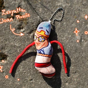 Könnte beinhalten: Ein Plüschwurm-Spielzeug mit der Aufschrift "Zenyatta Worm Plushie" mit einem Cartoon-Design. Der Wurm hat einen blau-gelben Hut, eine Brille und rote Arme. Es ist an einer silbernen Kette befestigt.