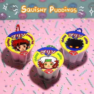 May include: Three small, clear plastic pudding pots with colourful lids. Each lid features a cartoon character and Japanese text. The text "Squishy Puddings" is above the pots. The background is pink with a repeating pattern of smiley faces.