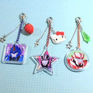 Op de afbeelding: Drie acryl sleutelhangers met metalen karabijnsluitingen en zilveren kettinkjes. De eerste is een vierkant met een blauwe robot, een rode aardbei-bedel en een klein sterretje. De tweede is stervormig met een paarse robot en een Hello Kitty-bedel. De derde is een cirkel met een rood-witte robot en een groene kristal-bedel.