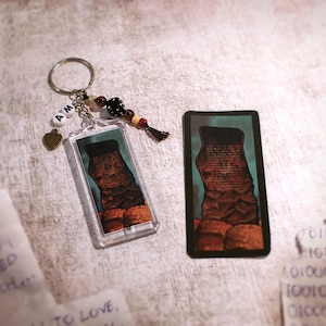 May include: A rectangular keychain featuring a dark, rocky monolith illustration, attached to a metal ring with white letter beads spelling AM, a small bronze heart charm, and a black and brown beaded tassel. A matching rectangular sticker is included.
