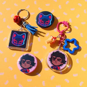 Puede incluir: Un conjunto de accesorios que incluye dos chapas redondas, un llavero cuadrado de acrílico con una borla azul y un cascabel rojo, y un llavero circular de acrílico con un cierre rosa en forma de estrella y un dije de helado.