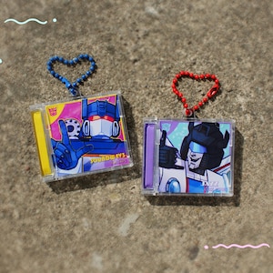 Può includere: Due portachiavi quadrati in acrilico con illustrazioni di personaggi. Il portachiavi sinistro ha una catena blu a forma di cuore e raffigura un robot blu con il testo SOUNDWAVE. Quello destro ha una catena rossa e raffigura un robot bianco con il testo JAZZ.