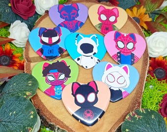 May include: Nine heart-shaped pins featuring different Spider-Man characters from the Spider-Verse movie. The pins are on a wooden surface with green moss and flowers.