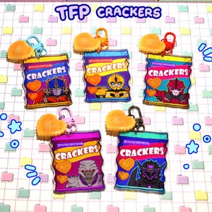 Peut inclure: Cinq breloques acryliques Transformers Cracker avec des crackers en forme de cœur. Chaque breloque présente un design de personnage différent sur un sac de crackers, avec le texte "CRACKERS". Le fond est une grille de carrés aux couleurs pastel.