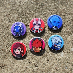 Peut inclure: Un ensemble de six badges ronds illustrés de personnages stylisés et colorés. Les motifs incluent une silhouette à capuche violette, un extraterrestre rose, un personnage à la peau bleue, une personne aux cheveux roux, une créature rouge et un alien bleu.