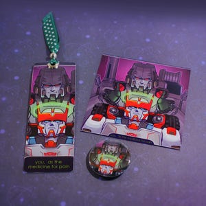 Puede incluir: Un marcapáginas rectangular con una cinta verde de lunares, una impresión cuadrada y un pin circular, todos con una ilustración gráfica de dos robots con el texto "you, as the medicine for pain" impreso en el marcapáginas y la impresión.