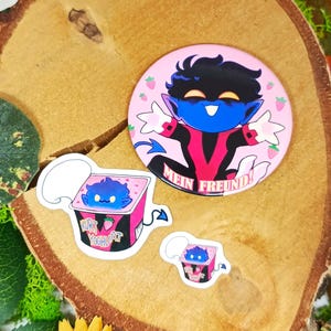 Può includere: Una collezione di adesivi e un bottone con un personaggio dei cartoni animati blu con un abito rosso e nero. Il bottone ha la scritta "MEIN FREUND!" ed è circondato da fragole. Gli adesivi includono il testo "KURT YOGHURT".