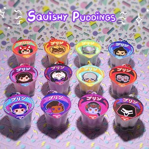 May include: An assortment of colorful, small pudding cups with character illustrations on the lids. The text "Squishy Puddings" is displayed above the cups. The cups are arranged on a patterned surface with a variety of colors.