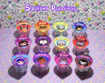 May include: An assortment of colorful, small pudding cups with character illustrations on the lids. The text "Squishy Puddings" is displayed above the cups. The cups are arranged on a patterned surface with a variety of colors.