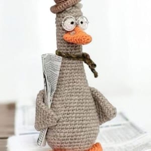 May include: A crocheted goose figurine wearing a brown hat, glasses, and a scarf, holding a newspaper. The goose has an orange beak and feet. The figurine is grey and sits on a stack of newspapers.