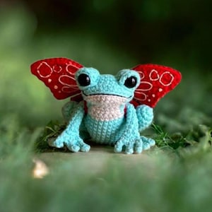 May include: A crocheted frog with light blue body, white chest, and large black eyes. It has red wings with white embroidered details. The frog is sitting on a surface with green foliage in the background.