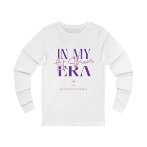 Puede incluir: Camiseta blanca de manga larga con el texto "IN MY ERA" en morado y rosa. La camiseta también presenta el texto "Let Them" en cursiva y la frase "LIVETHEBEAUTIFULNESS" en la parte inferior.