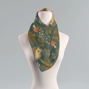 May include: A teal scarf with a woodland pattern featuring hedgehogs, mushrooms, pinecones, and sprigs. The scarf is displayed on a mannequin, showcasing the triangular shape and the detailed print. The design has a vintage, hand-drawn aesthetic.