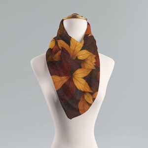 May include: A scarf with a leaf pattern in shades of brown, orange, and yellow is displayed on a mannequin. The scarf has a triangular shape and is made of a soft fabric. The leaves are arranged in a repeating pattern.