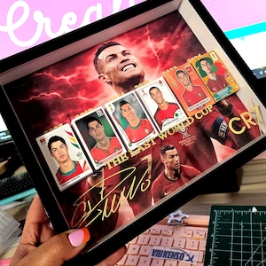 Puede incluir: Un marco que muestra un gran retrato de Cristiano Ronaldo sobre un fondo rojo con rayos. Incluye cinco tarjetas coleccionables más pequeñas del atleta con varias equipaciones de fútbol, el texto THE LAST WORLD CUP y una firma dorada.