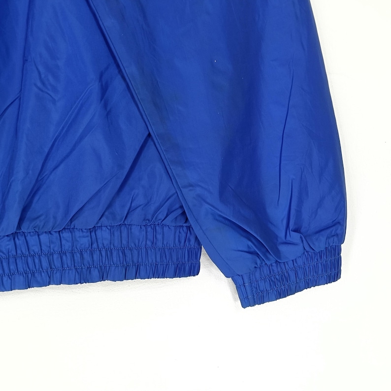 May include: A close-up of a royal blue jacket. The jacket has a gathered waist and cuffs, and a split front. The fabric appears to be a lightweight, possibly nylon or polyester material. The jacket is a solid color with no visible patterns.