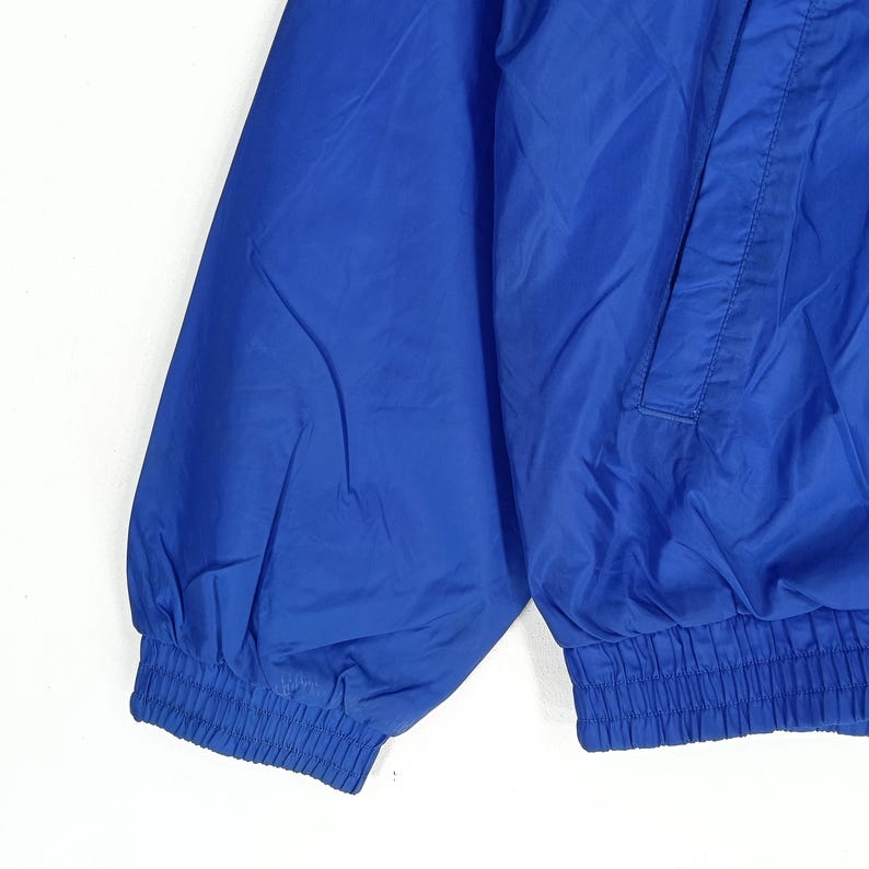 May include: Close-up of a bright blue jacket sleeve. The jacket has a ribbed cuff and a pocket detail. The fabric appears to be a lightweight, possibly nylon or polyester material. The jacket is a solid, vibrant blue color.