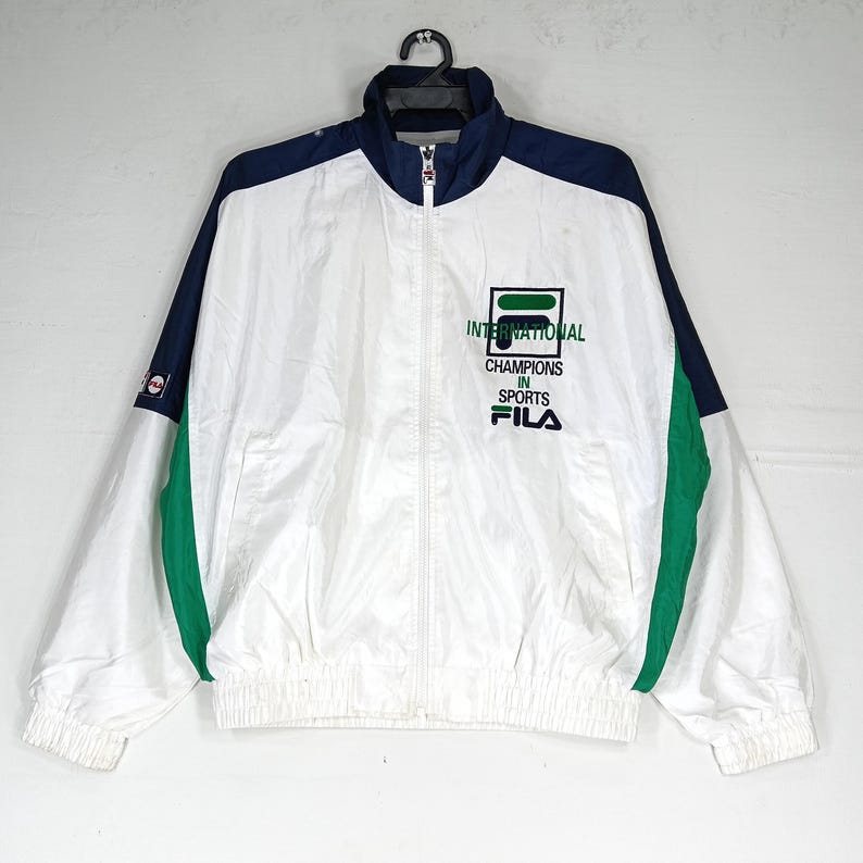 May include: White, navy blue, and green FILA track jacket with a full zipper closure. The jacket features the text "INTERNATIONAL CHAMPIONS IN SPORTS FILA" on the chest. The jacket has a stand-up collar and elastic cuffs and hem.