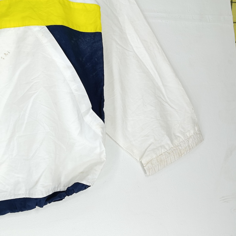 May include: White, yellow, and navy blue windbreaker jacket. The jacket features a color block design with a yellow stripe across the top, a navy blue angled panel, and a white body. The sleeve is visible on the right side.