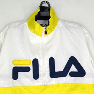 May include: White Fila track jacket with a yellow collar and hem. The jacket features a full zipper and the Fila logo in navy blue and yellow on the front. The jacket is hanging on a black hanger.