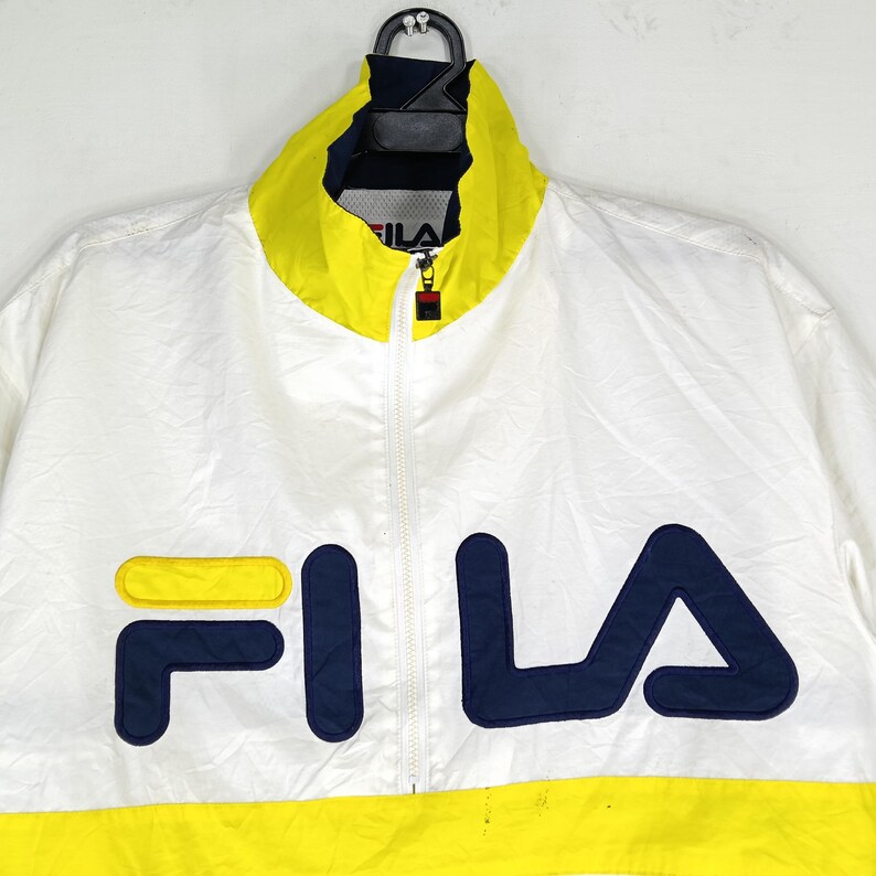 May include: White Fila track jacket with a yellow collar and hem. The jacket features a full zipper and the Fila logo in navy blue and yellow on the front. The jacket is hanging on a black hanger.