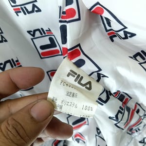 May include: Close-up of a white fabric with a repeating pattern of the FILA logo in red, navy, and black. A tag with the FILA logo and Japanese text is visible. The fabric appears to be part of a jacket or sportswear.