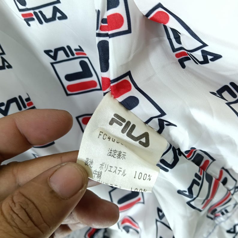 May include: Close-up of a white fabric with a repeating pattern of the FILA logo in red, navy, and black. A tag with the FILA logo and Japanese text is visible. The fabric appears to be part of a jacket or sportswear.