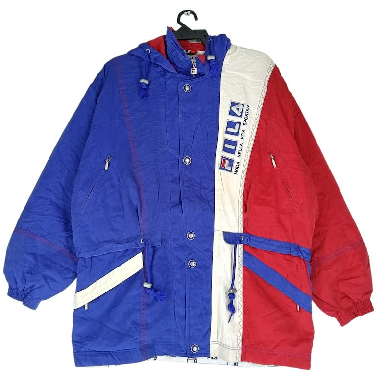 May include: A vintage Fila jacket in blue, red, and white. The jacket features a hood, multiple pockets, and the Fila logo. The design includes a color-blocked pattern with the text "Moda Nella Vita Sportiva".