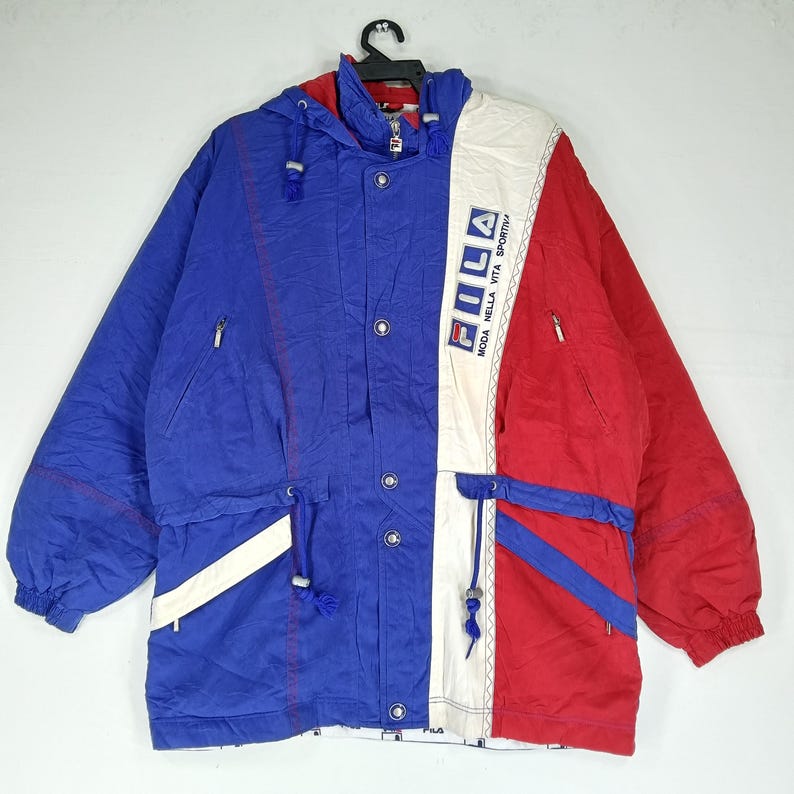 May include: A vintage Fila jacket in blue, red, and white. The jacket features a hood, multiple pockets, and the Fila logo. The design includes a drawstring waist and a zipper closure. The jacket is a retro style.