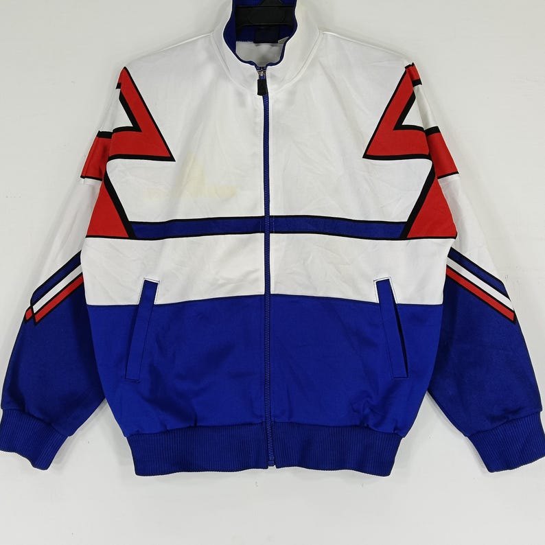 May include: A white and blue track jacket with red and black geometric designs. The jacket has a zipper closure and a stand-up collar.