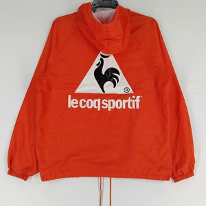 K&ouml;nnte beinhalten: Leuchtend orangefarbene Kapuzenjacke mit dem Le Coq Sportif Logo auf dem R&uuml;cken. Das Logo zeigt einen schwarzen Hahn in einem wei&szlig;en Dreieck, darunter der Markenname in wei&szlig;er Schrift. Die Jacke hat einen Kordelzug am Saum.