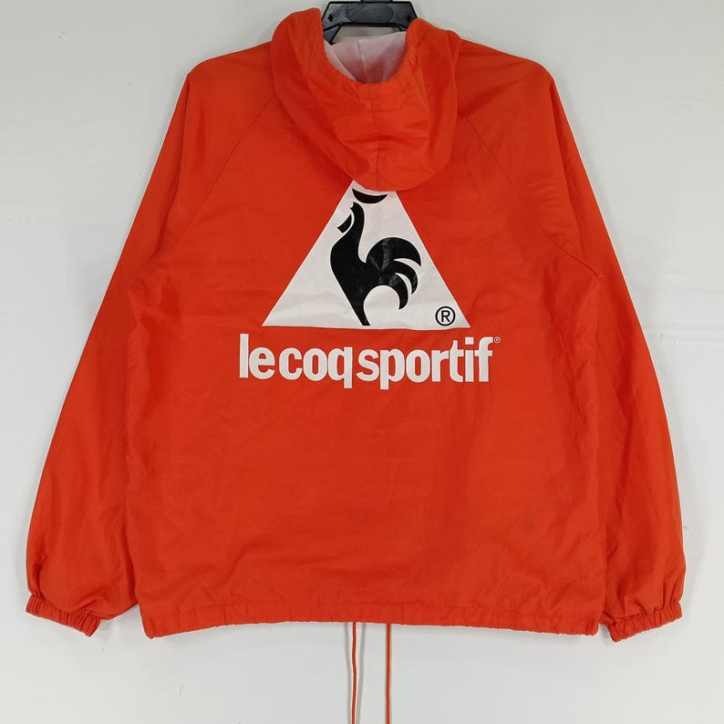K&ouml;nnte beinhalten: Leuchtend orangefarbene Kapuzenjacke mit dem Le Coq Sportif Logo auf dem R&uuml;cken. Das Logo zeigt einen schwarzen Hahn in einem wei&szlig;en Dreieck, darunter der Markenname in wei&szlig;er Schrift. Die Jacke hat einen Kordelzug am Saum.