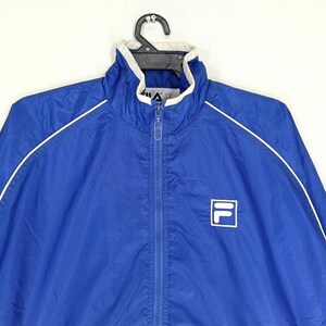 May include: A blue Fila track jacket with a white collar and white trim along the shoulders and sleeves. The jacket has a full zipper and the Fila logo on the chest.