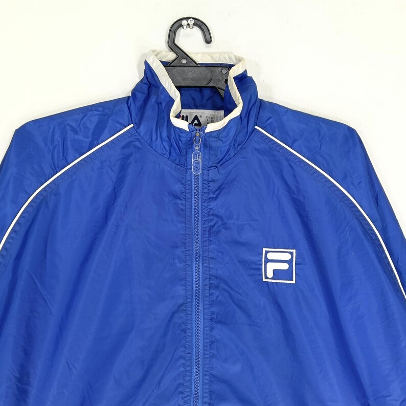 May include: A blue Fila track jacket with a white collar and white trim along the shoulders and sleeves. The jacket has a full zipper and the Fila logo on the chest.