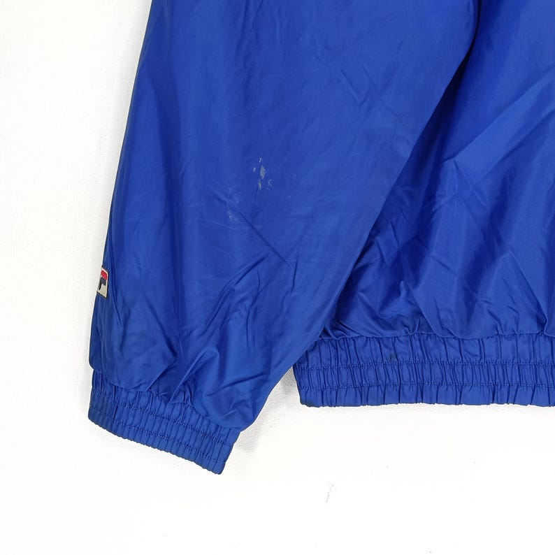 May include: A close-up of a blue jacket sleeve. The jacket has a ribbed elastic cuff and a small logo on the sleeve. The jacket appears to be made of a lightweight, possibly nylon, material. There are a few small stains on the jacket.