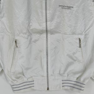 May include: White Courr&egrave;ges sport futur zip-up jacket. The jacket has a shiny, textured appearance with a ribbed hem and cuffs. The Courr&egrave;ges logo is printed in black on the upper right chest.