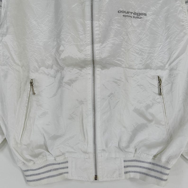 May include: White Courr&egrave;ges sport futur zip-up jacket. The jacket has a shiny, textured appearance with a ribbed hem and cuffs. The Courr&egrave;ges logo is printed in black on the upper right chest.