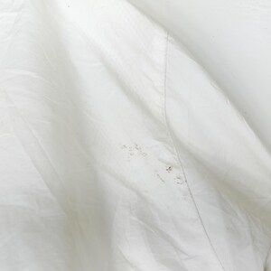 May include: Close-up of a white fabric with a subtle textured pattern. The fabric is slightly wrinkled, with a visible seam running diagonally across the image. There are small, dark spots on the fabric.