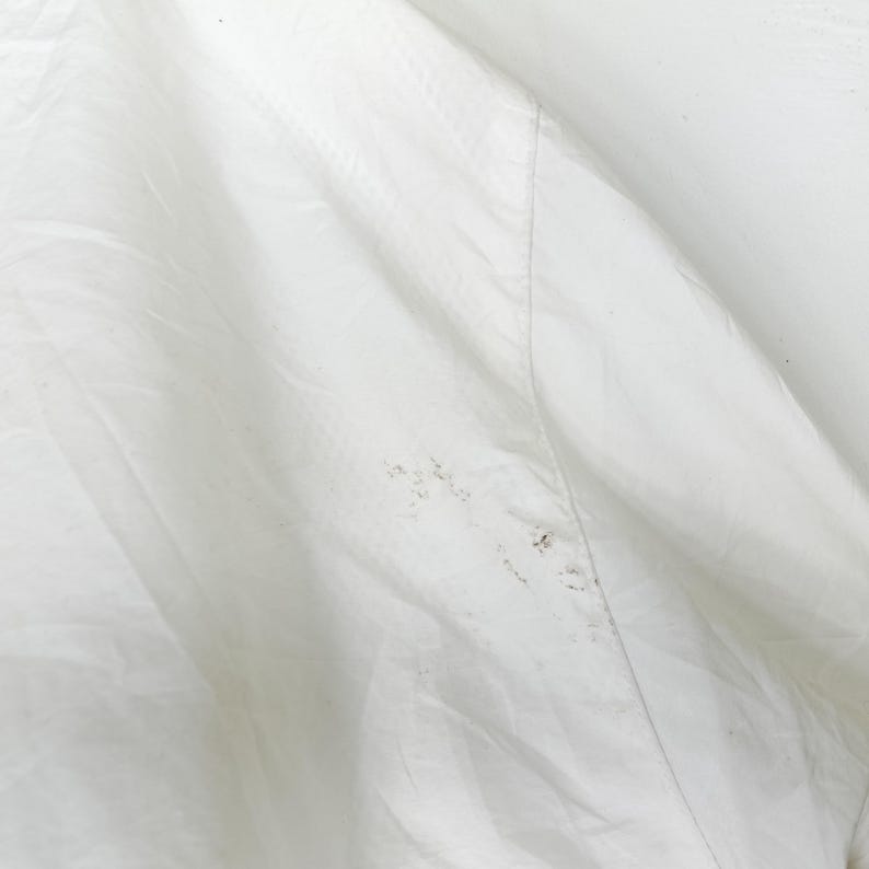 May include: Close-up of a white fabric with a subtle textured pattern. The fabric is slightly wrinkled, with a visible seam running diagonally across the image. There are small, dark spots on the fabric.