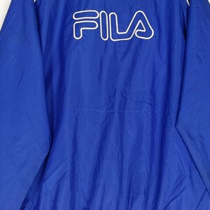 May include: A blue jacket with the white FILA logo across the chest. The jacket has a white trim along the collar and zipper. The material appears to be a lightweight nylon or polyester.