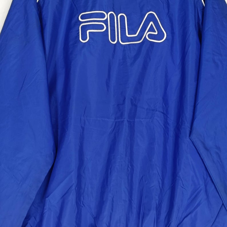 May include: A blue jacket with the white FILA logo across the chest. The jacket has a white trim along the collar and zipper. The material appears to be a lightweight nylon or polyester.