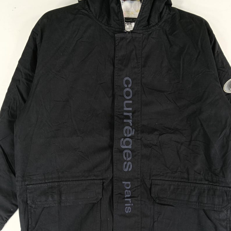 May include: Black hooded jacket with the text "courr&egrave;ges paris" vertically down the front. The jacket has a front zipper, two front pockets, and a small logo on the upper right sleeve. The jacket is made of a durable material.
