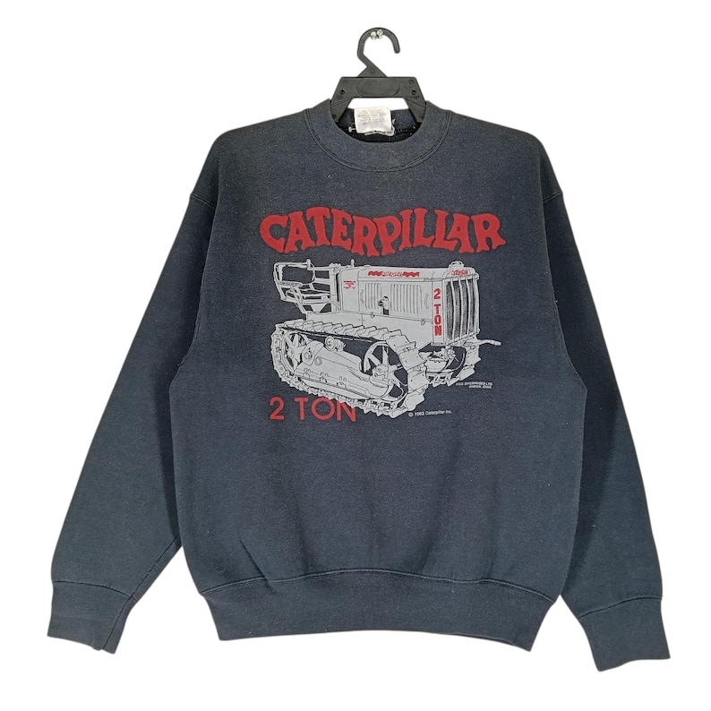 K&ouml;nnte beinhalten: Dunkelgraues Sweatshirt mit einem roten und grauen Caterpillar-Traktor-Motiv und dem Text "CATERPILLAR" und "2 TON". Das Sweatshirt h&auml;ngt an einem schwarzen Kleiderb&uuml;gel.
