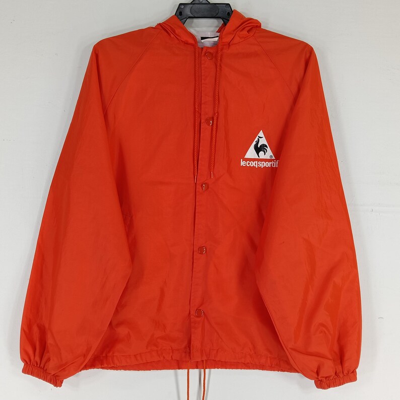 K&ouml;nnte beinhalten: Leuchtend orangefarbene Kapuzenjacke mit Druckknopfverschluss. Sie hat ein wei&szlig;es Logo mit einem Hahn-Emblem auf der Brust. Die Jacke hat einen Kordelzug am Saum und elastische B&uuml;ndchen. Ein l&auml;ssiges Oberbekleidungsst&uuml;ck.