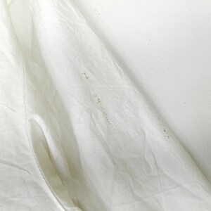 May include: Close-up of a white garment with visible folds and a pocket. The fabric appears to be slightly wrinkled, with several small brown stains scattered across the surface. The background is a plain white wall.