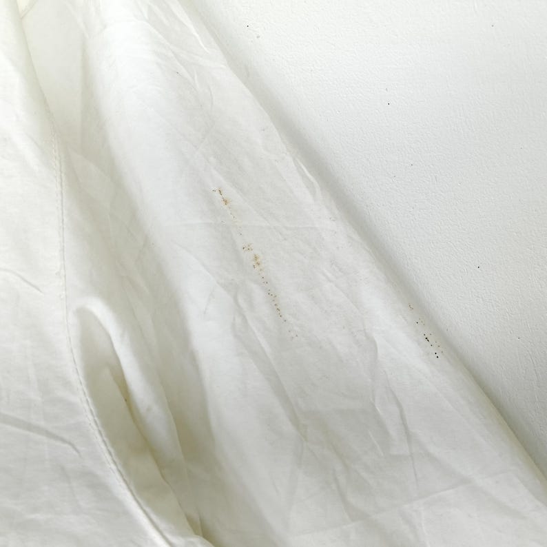 May include: Close-up of a white garment with visible folds and a pocket. The fabric appears to be slightly wrinkled, with several small brown stains scattered across the surface. The background is a plain white wall.