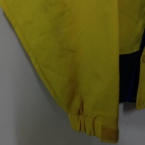 Puede incluir: Primer plano de una chaqueta amarilla con un panel azul y negro. La chaqueta tiene un pu&ntilde;o de velcro y cierre de bot&oacute;n. La tela parece ser un material ligero.
