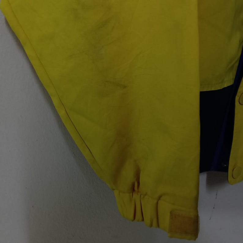 Puede incluir: Primer plano de una chaqueta amarilla con un panel azul y negro. La chaqueta tiene un pu&ntilde;o de velcro y cierre de bot&oacute;n. La tela parece ser un material ligero.