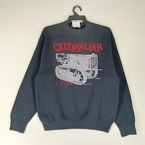 K&ouml;nnte beinhalten: Dunkelgraues Sweatshirt mit einem roten und grauen Caterpillar-Traktor-Motiv. Der Text "CATERPILLAR" steht dar&uuml;ber und "2 TON" darunter. Es hat einen Rundhalsausschnitt und lange &Auml;rmel.